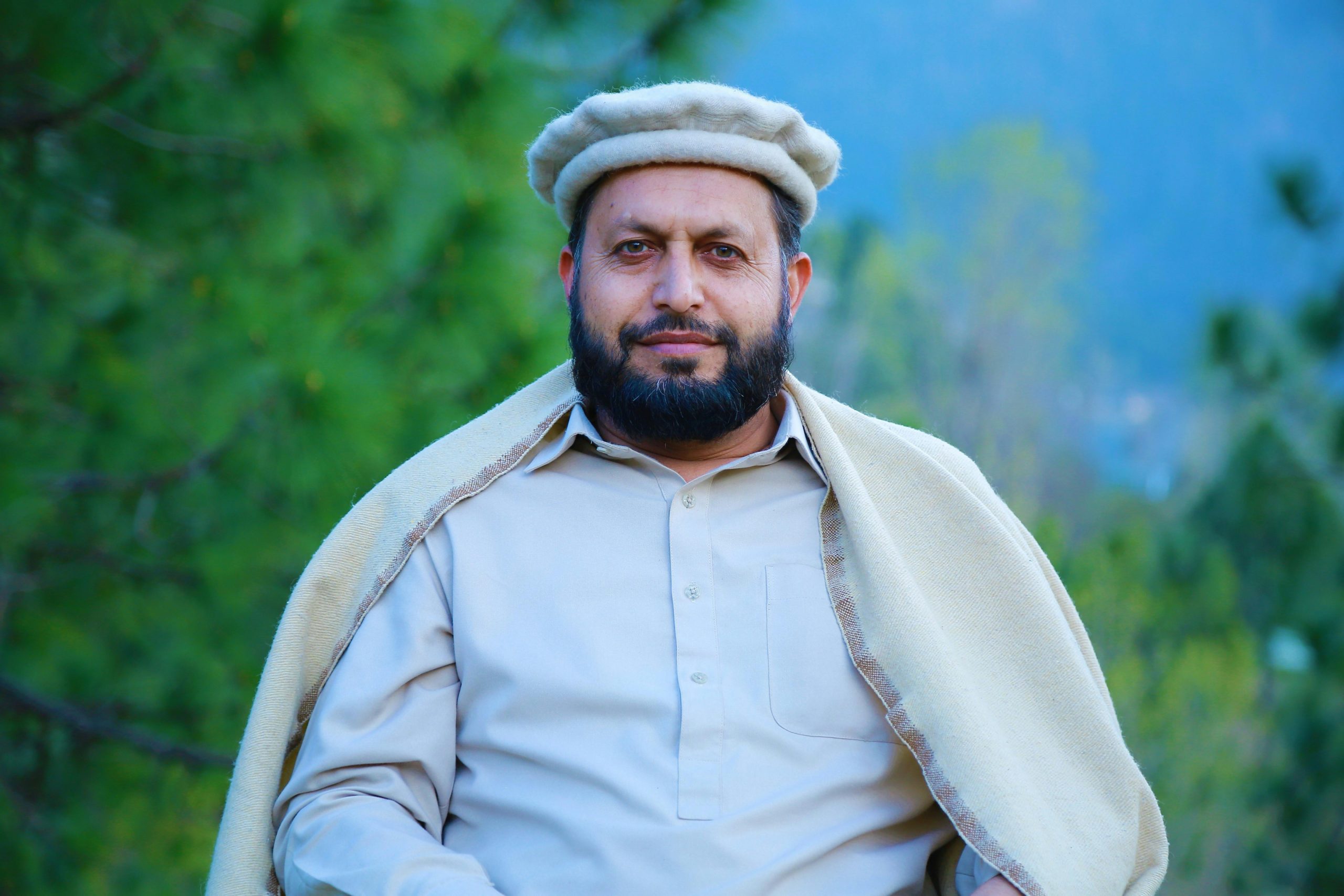 A man outdoors wearing a Chitrali cap, wrapped in a shawl and posing with a serene smile.