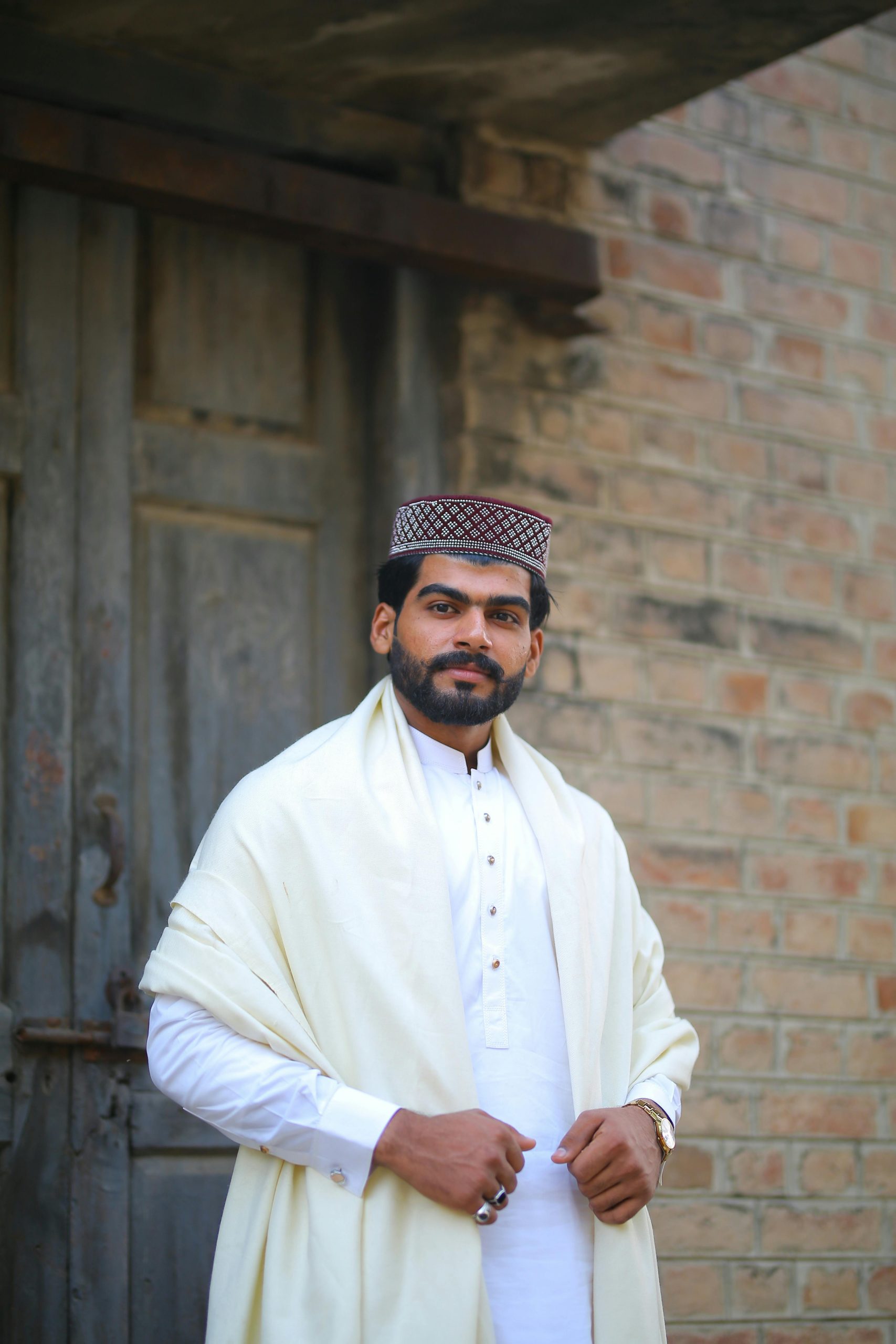 Portrait of a Pakistani man in traditional attire with a shawl and topi, standing outdoors.
